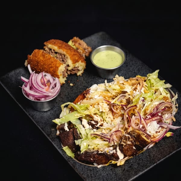 A black plate with a large taco, a side of red onions, a green dipping sauce, and two croquettes.