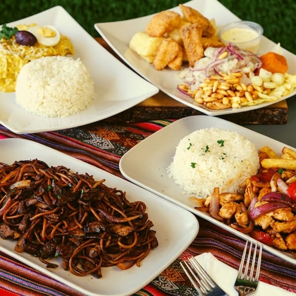 Four plates of Peruvian dishes, including lomo saltado, aji de gallina, and fried seafood, on a colorful striped tablecloth.