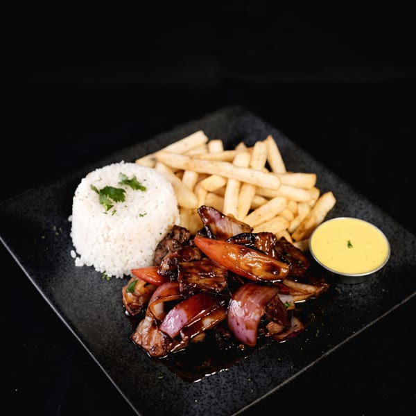 Lomo Saltado with white rice, french fries, and a side of yellow sauce on a black plate.