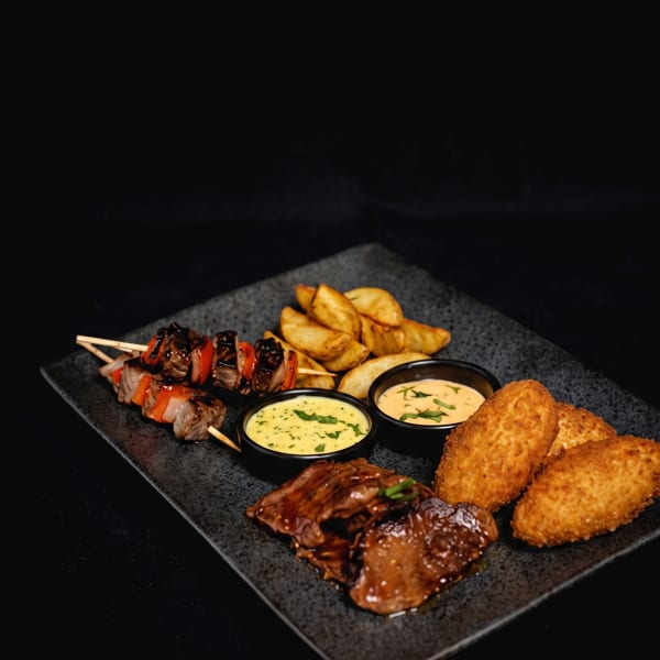 A black rectangular plate with beef skewers, potato wedges, fried croquettes, and two dipping sauces.